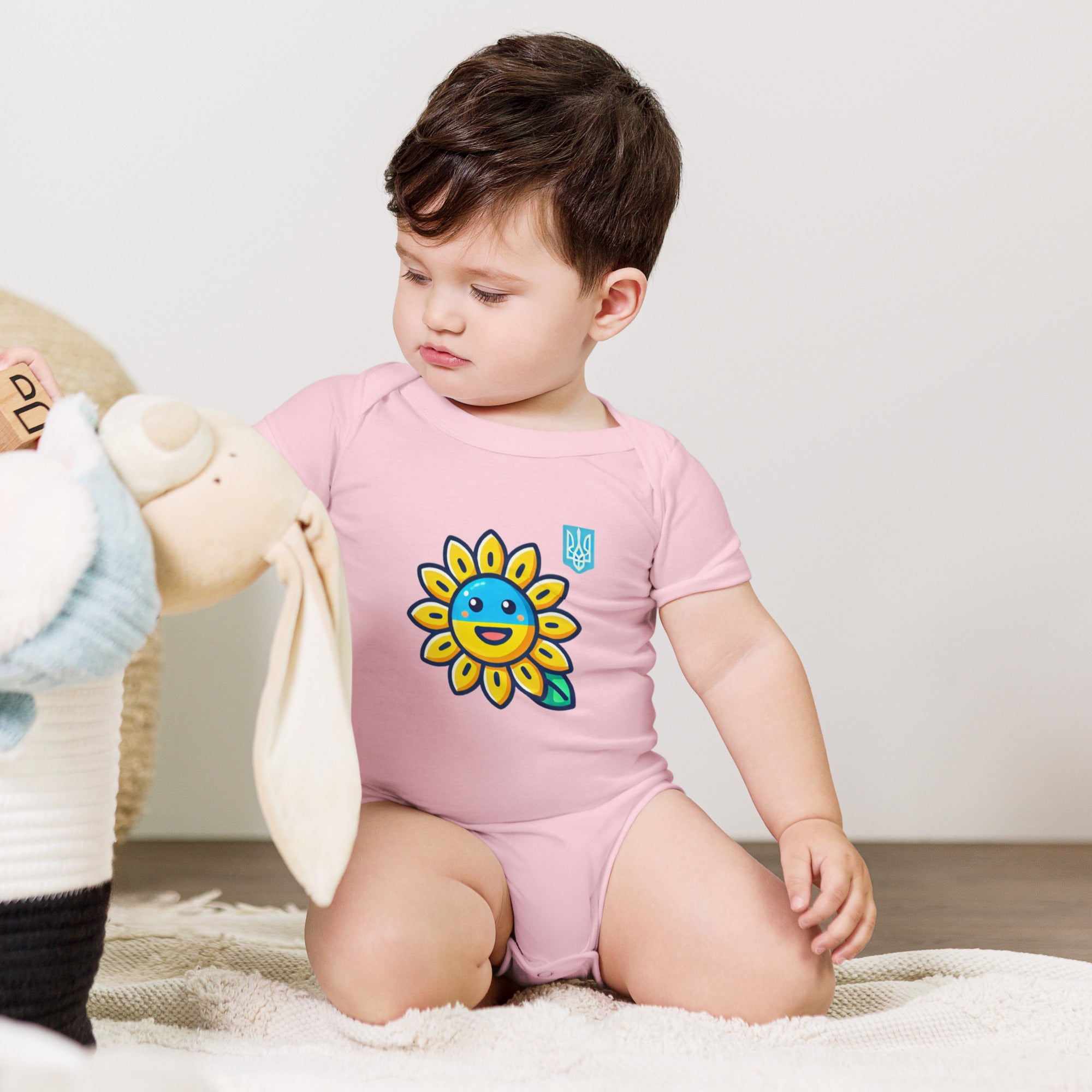 Baby wearing pink bodysuit with sunflower and Ukrainian flag design, sitting on the floor with toys nearby.