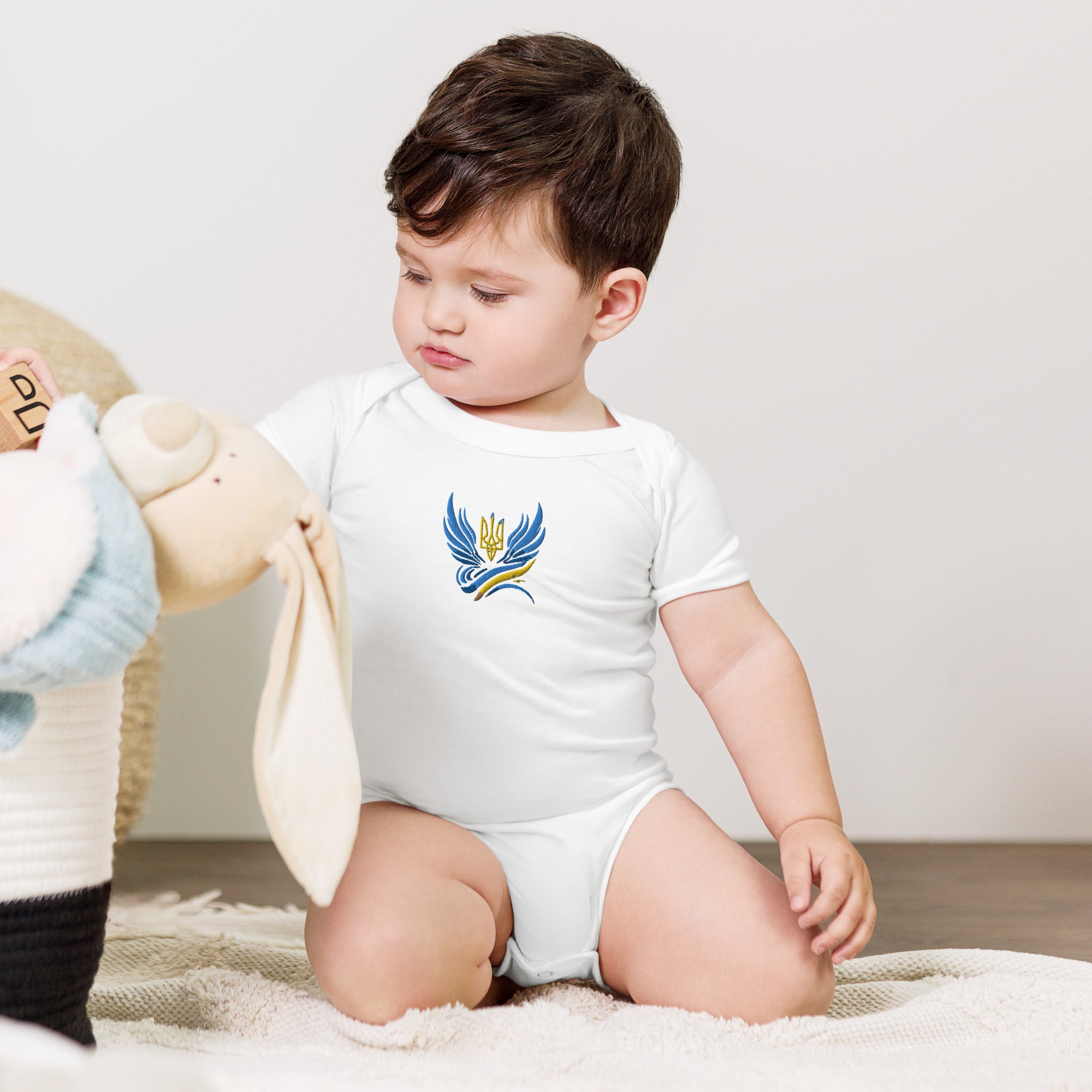 Baby in cotton one piece with Ukrainian Tryzub logo, sitting on a soft surface.