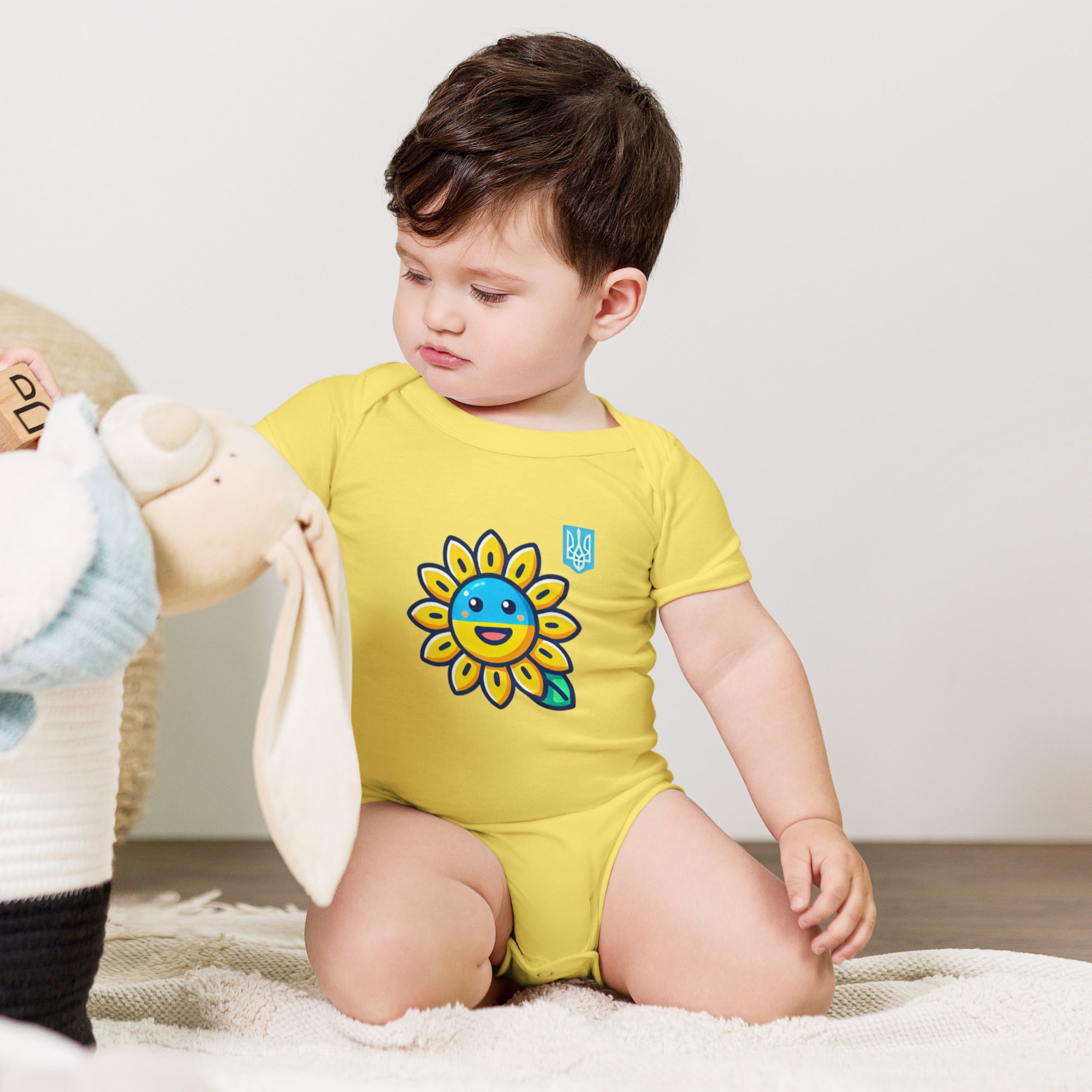 Baby wearing yellow bodysuit with sunflower and Ukrainian flag design, sitting near toys.