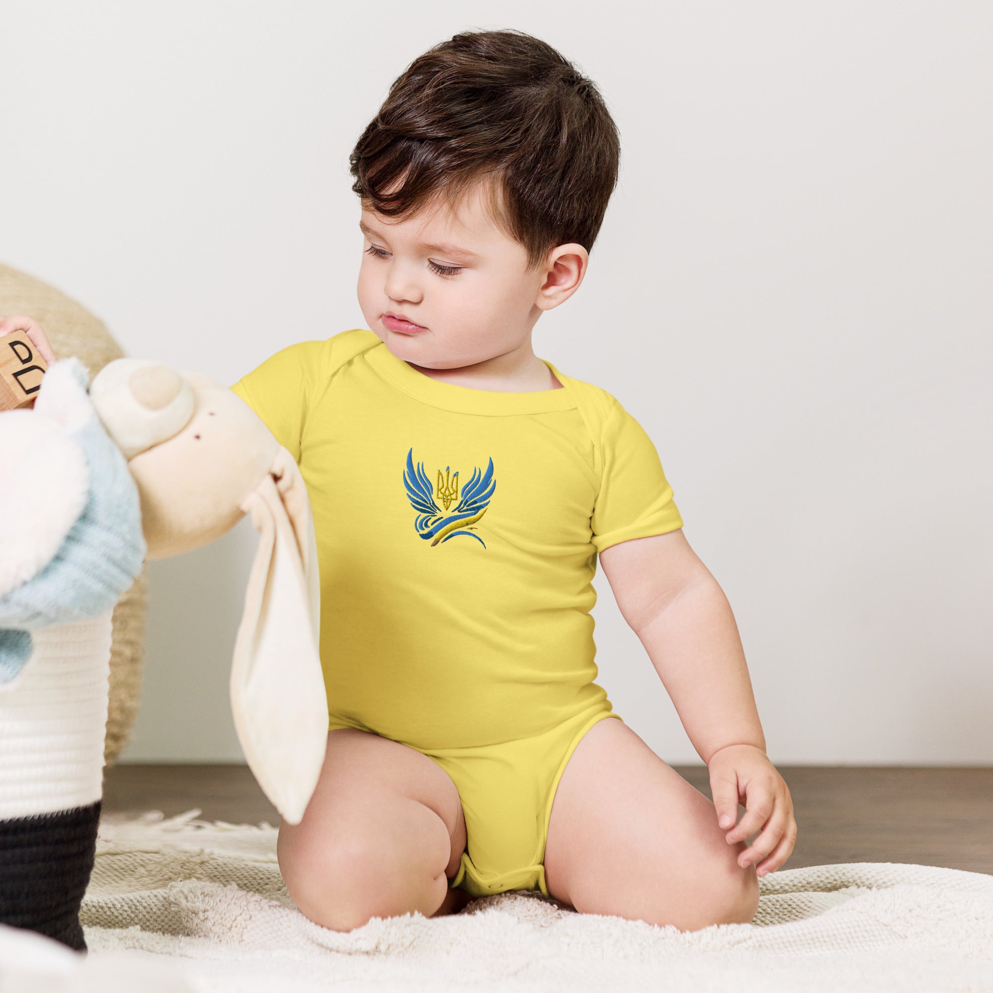 Baby wearing yellow short sleeve one piece with Ukrainian Tryzub logo, sitting on a soft rug next to a stuffed animal.