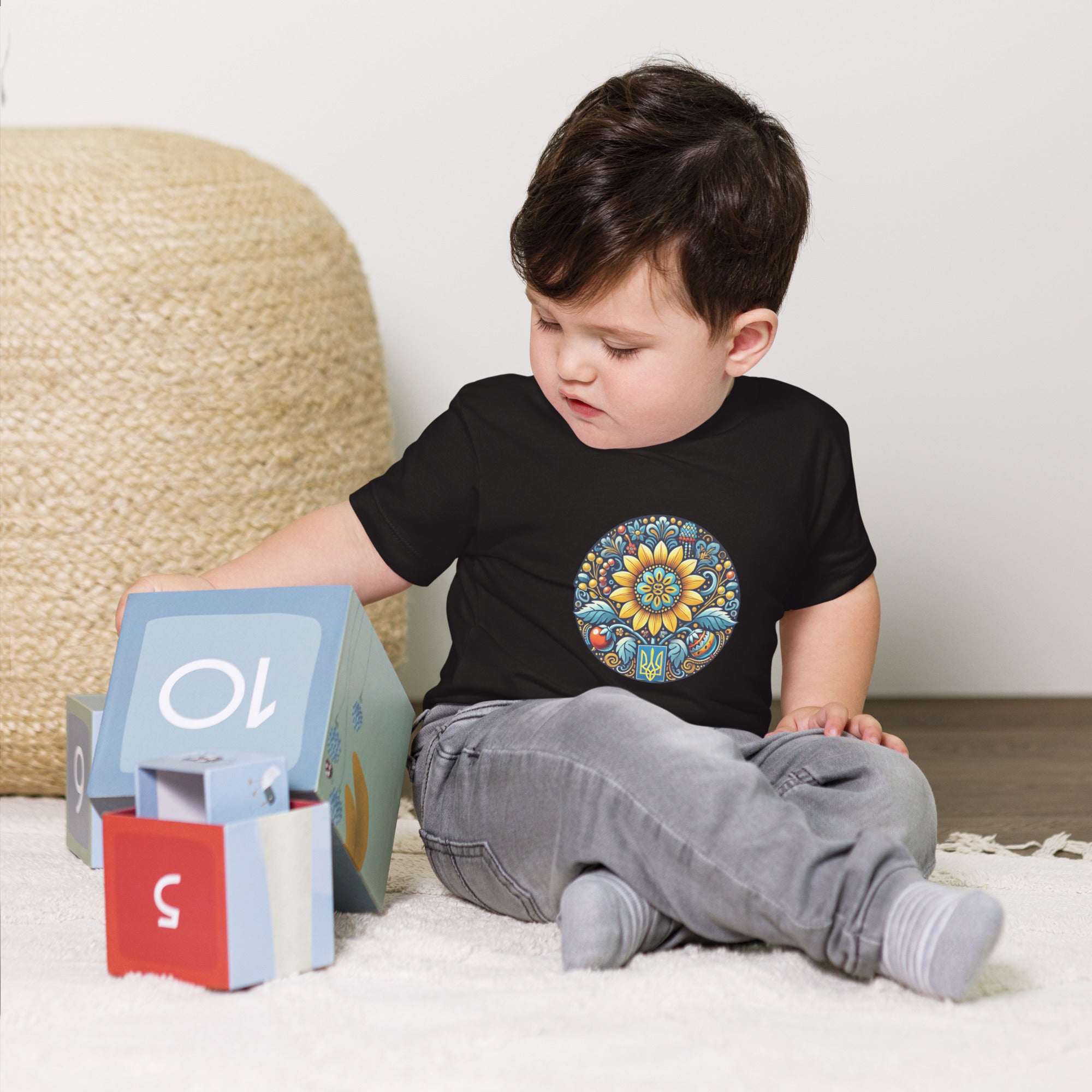 Toddler wearing a black baby jersey tee with colorful Ukrainian symbol, playing with blocks on a wooden floor.