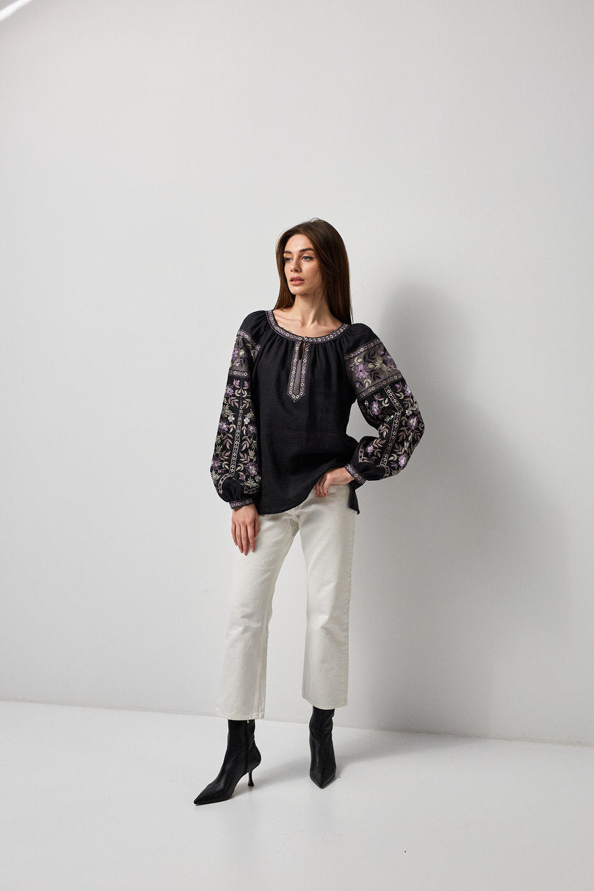 Woman wearing a black linen Ukrainian vyshyvanka blouse with embroidered sleeves and symbolic back ornament, paired with white pants.