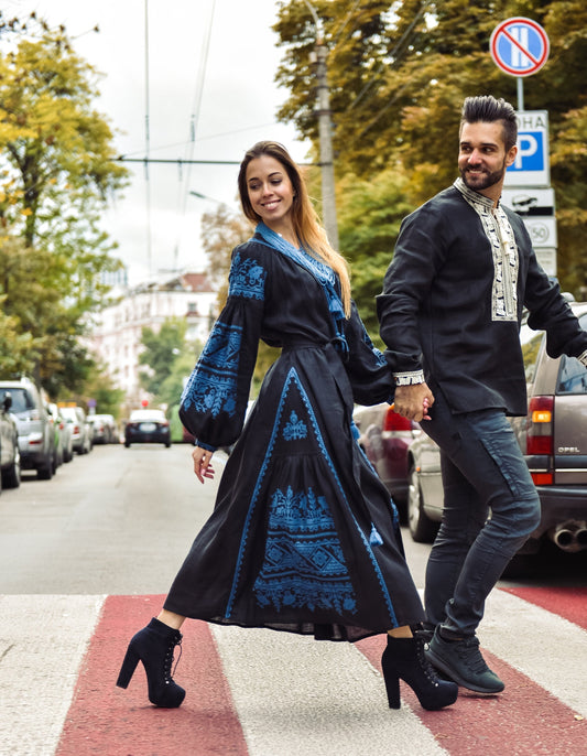 Couple wearing black embroidered Ukrainian Vibe vyshyvanka, walking on street; woman's boho-style linen kaftan dress with cultural motifs.