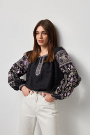Woman wearing black linen embroidered blouse with Ukrainian vyshyvanka design, featuring traditional symbolism and modern aesthetics.