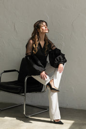 Model wearing black-on-black embroidered blouse with tulle inserts, seated on chair, showcasing Boho ethnic style.