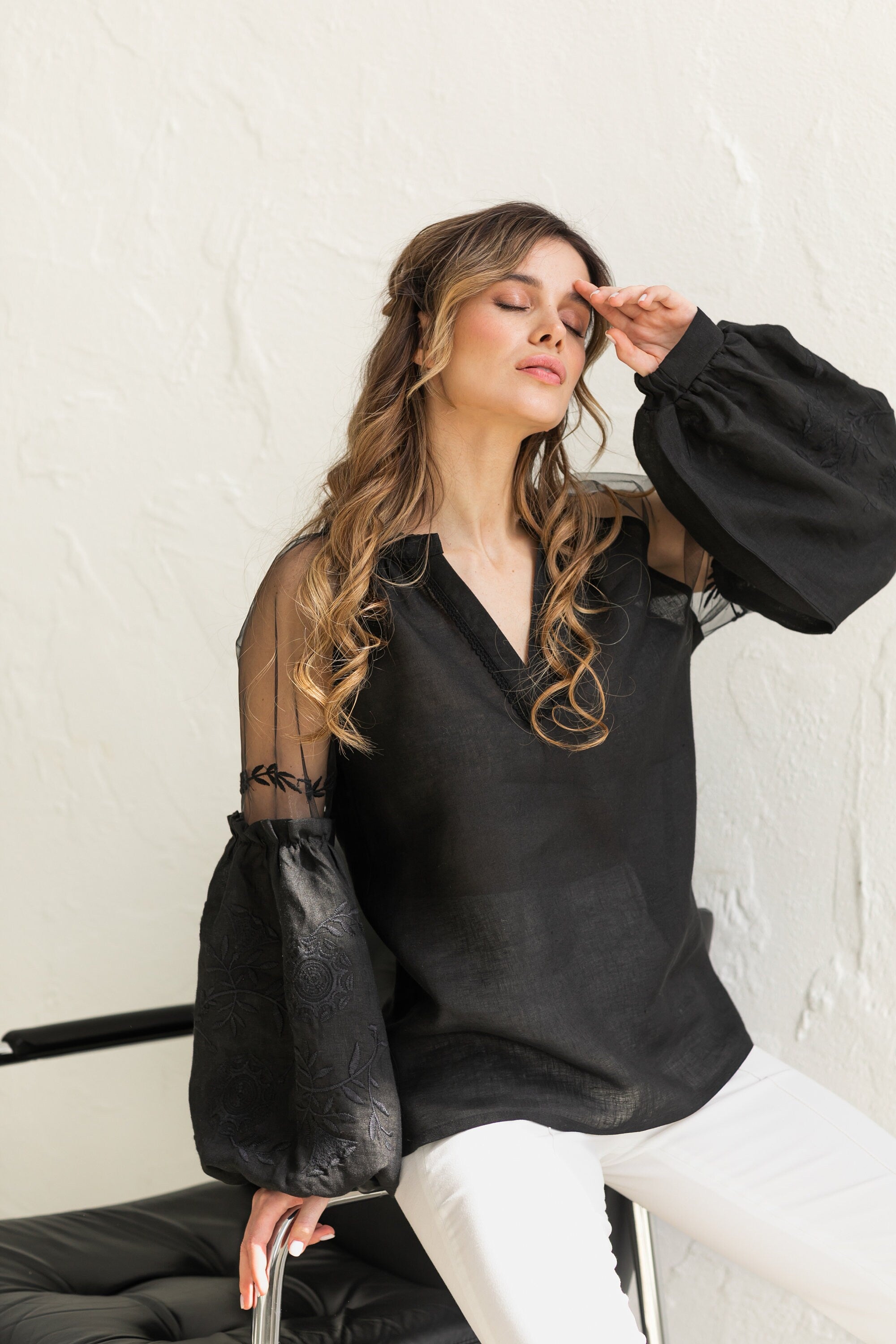 Woman wearing black-on-black Ukrainian embroidered blouse with tulle shoulder inserts, boho ethnic style, sitting elegantly on a chair.