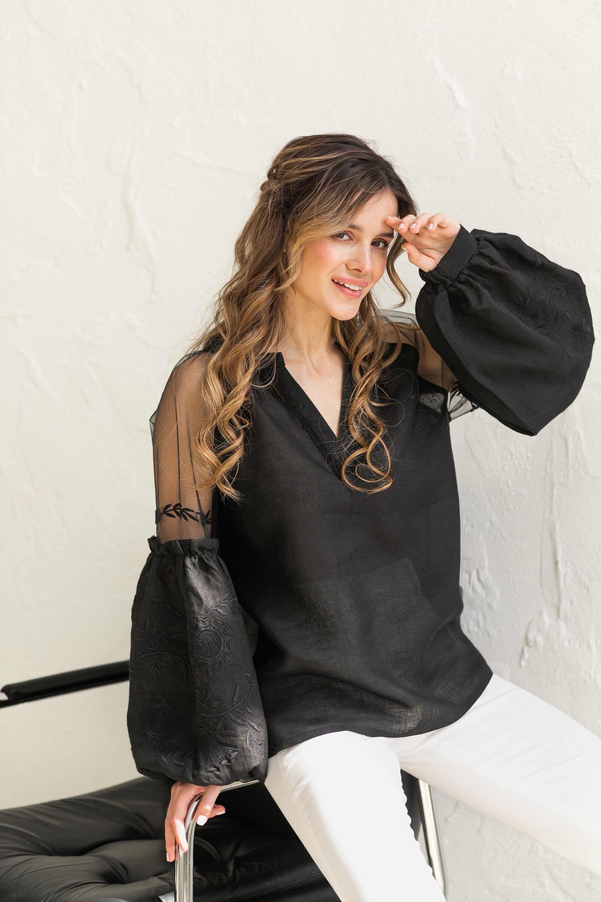Woman wearing black-on-black embroidered Ukrainian blouse with tulle shoulder inserts in boho ethnic style, seated on a black chair.