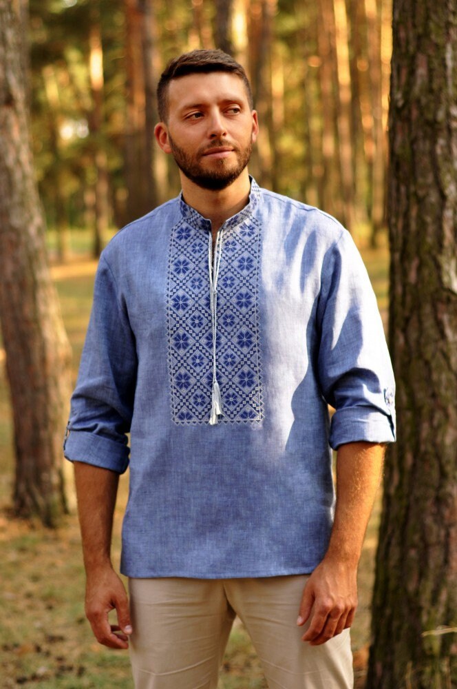 Man wearing blue embroidered Ukrainian Vyshyvanka linen shirt with Alatyr sign, standing in forest.