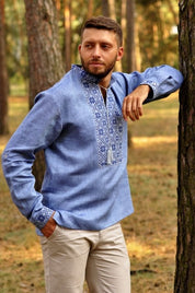 Man wearing blue Ukrainian embroidered linen shirt with Slavic Alatyr symbol in a forest setting.