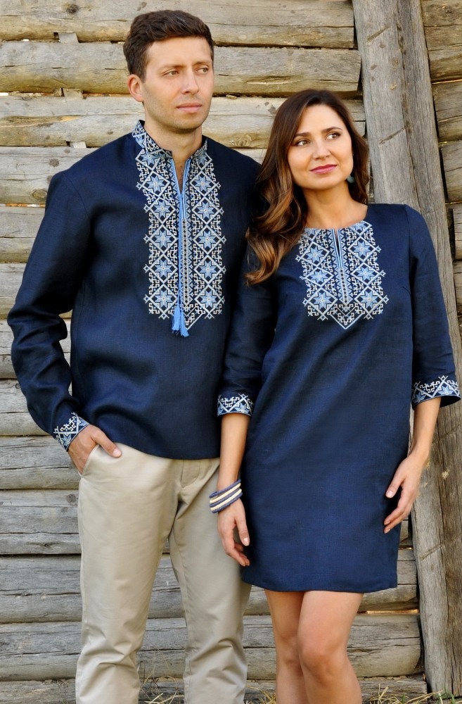Man and woman in traditional Ukrainian outfits with blue embroidery, standing in front of a wooden wall.