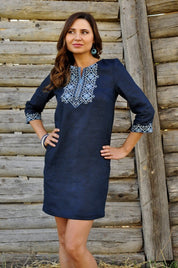 Woman wearing a blue linen embroidered mini dress with Ukrainian Vyshyvanka and Alatyr symbol, standing against a wooden fence.