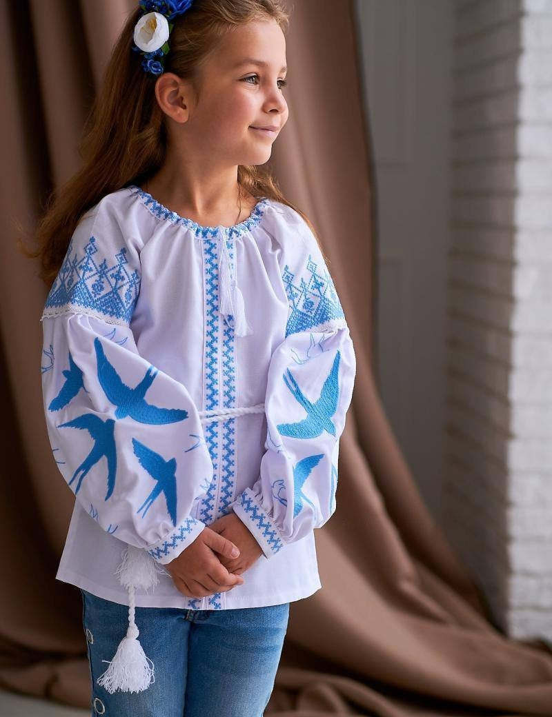 Girl wearing a traditional Ukrainian blouse with blue and yellow embroidery featuring swallows, symbolizing peace and freedom.