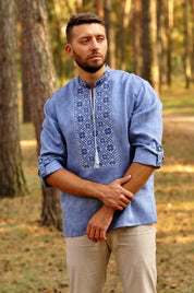 Man wearing blue embroidered Ukrainian Vyshyvanka linen shirt with Slavic symbolism, standing in a forest setting.