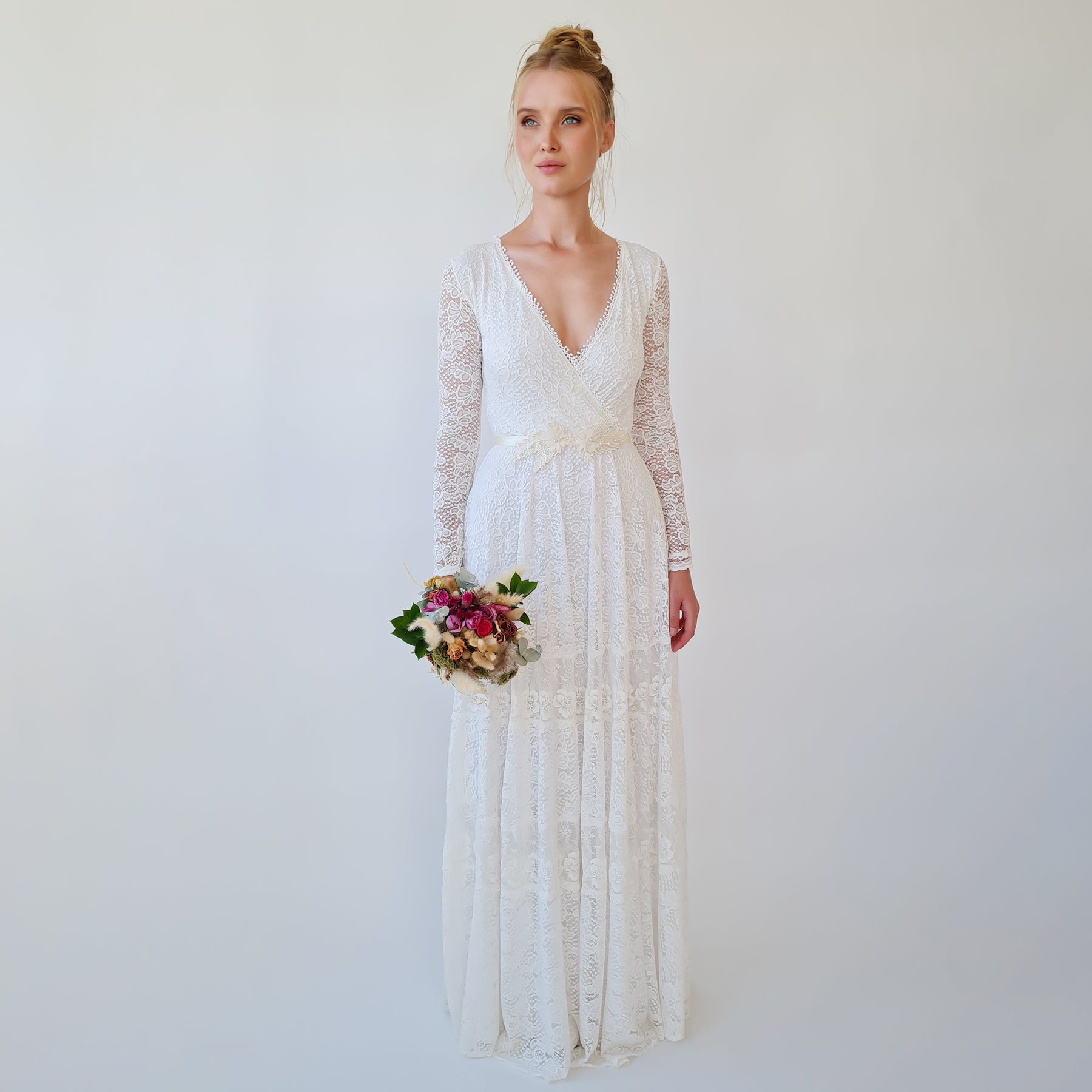 Bride in white gown wearing Isara Beaded Floral Sash with lace details and floral accents, holding a bouquet, standing against a plain background.