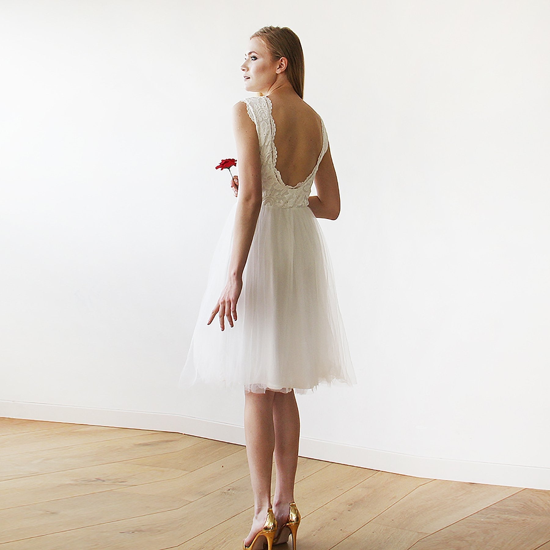Woman wearing Evarelle Midi Lace Tulle Wedding Dress with open-back design standing on wooden floor holding red flower