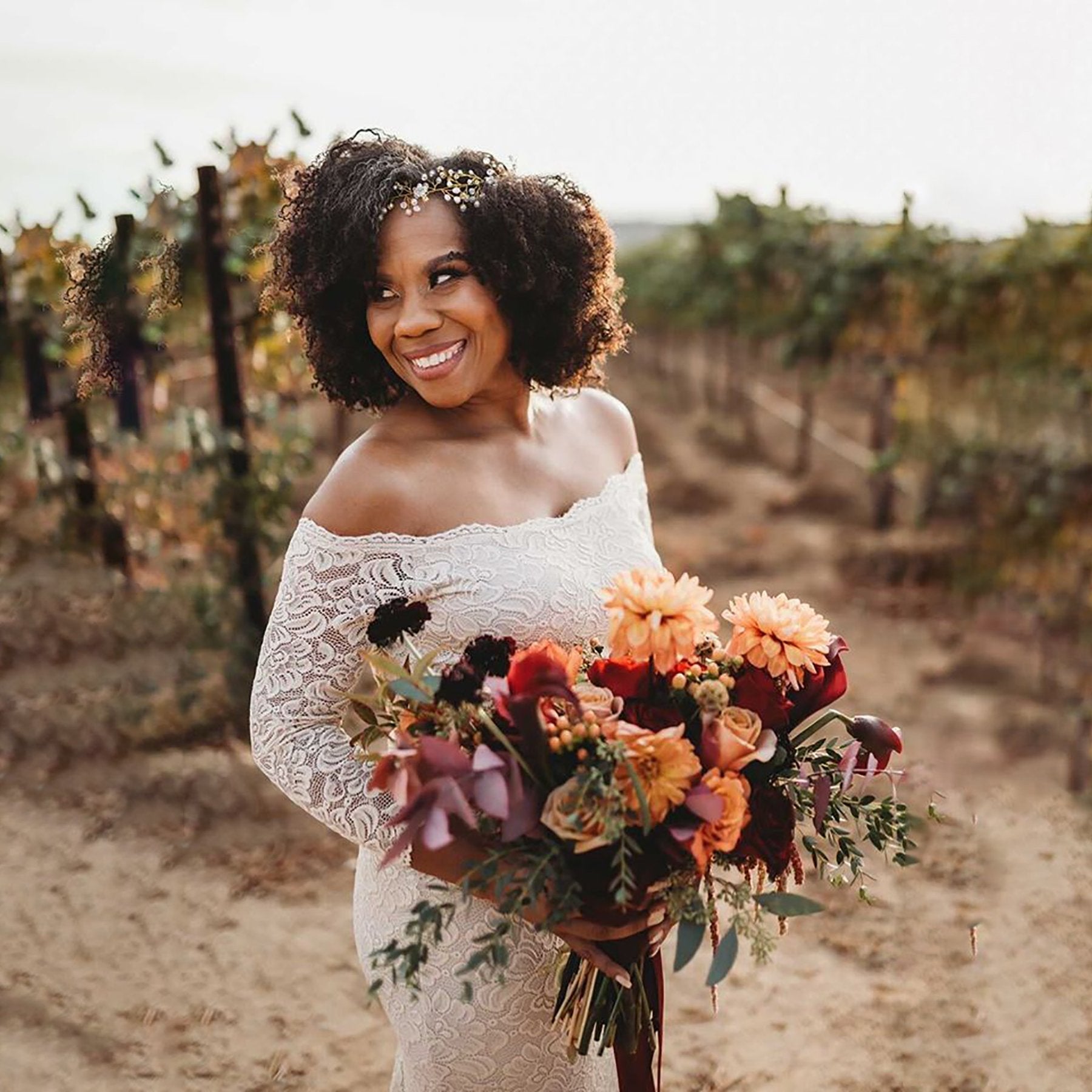 Bride in Mirelline champagne off-shoulder wedding dress holding vibrant bouquet, posing in vineyard with elegant off-the-shoulder lace design.