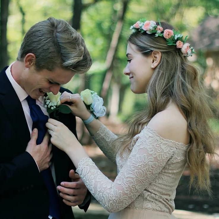 Woman in lace dress adjusts man's tie in outdoor setting, both smiling, with flowers in her hair, capturing a joyful moment.