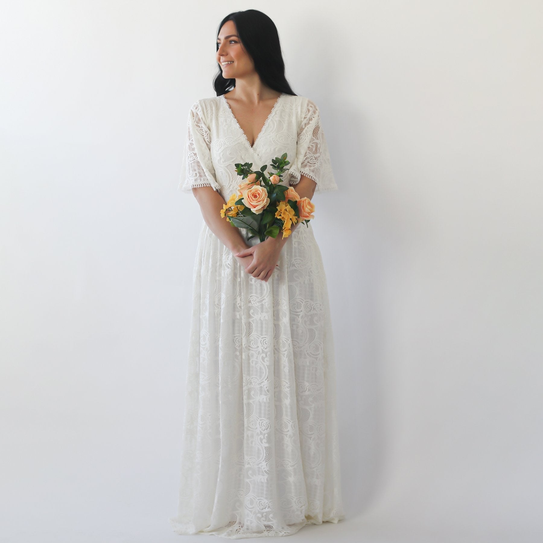 Woman wearing boho lace gown with butterfly sleeves and pockets, holding flowers. Ideal for bohemian weddings and sunlit ceremonies.