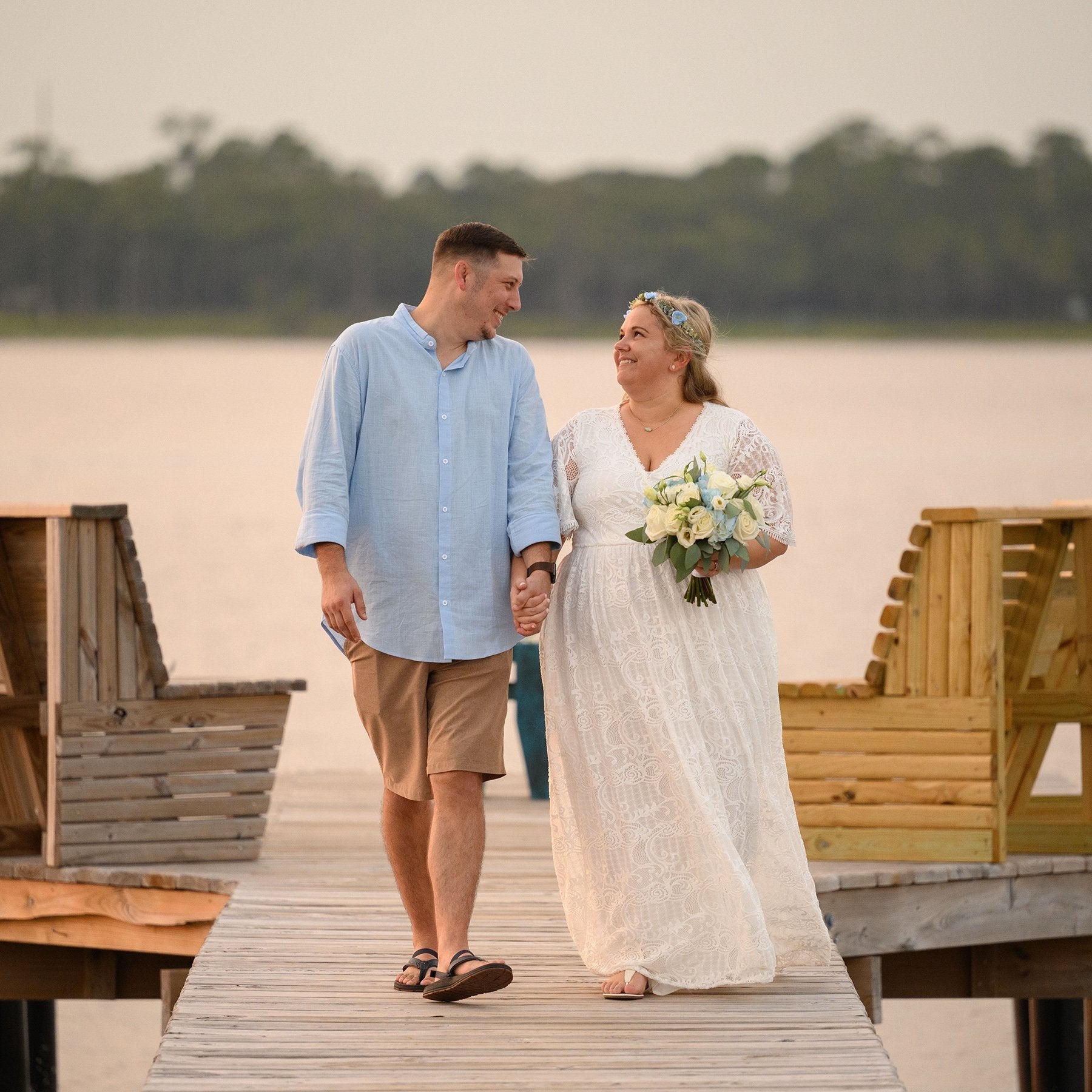 Couple walking on a dock; woman wearing the Marvelle Boho Lace Gown with butterfly sleeves, holding a bouquet, at sunset.