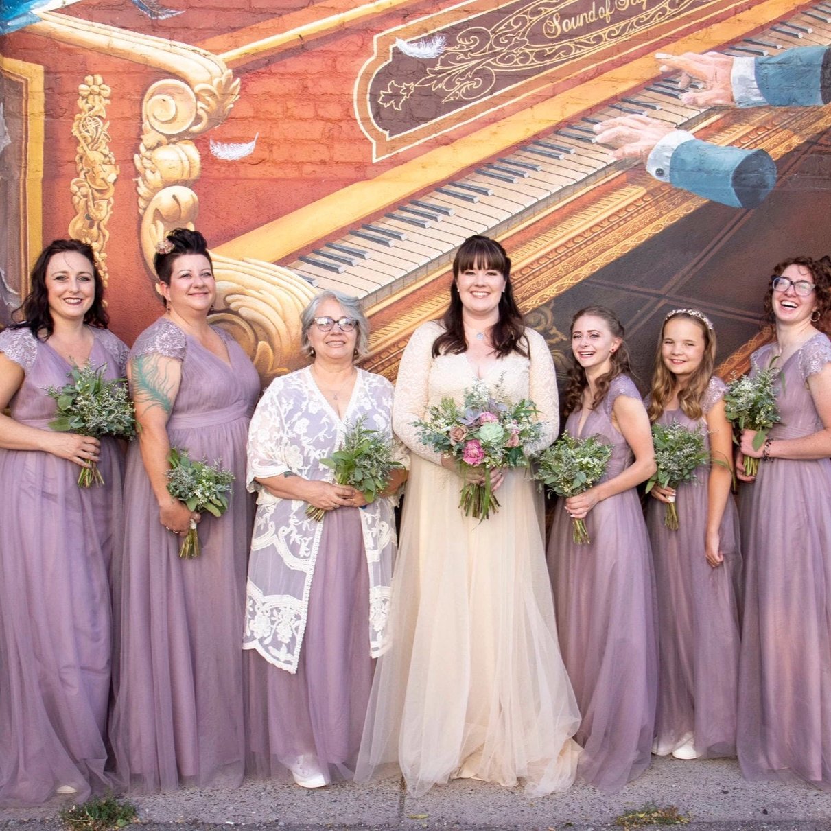 Bride in champagne lace gown with bridesmaids in lavender dresses holding bouquets, standing in front of a vibrant mural.