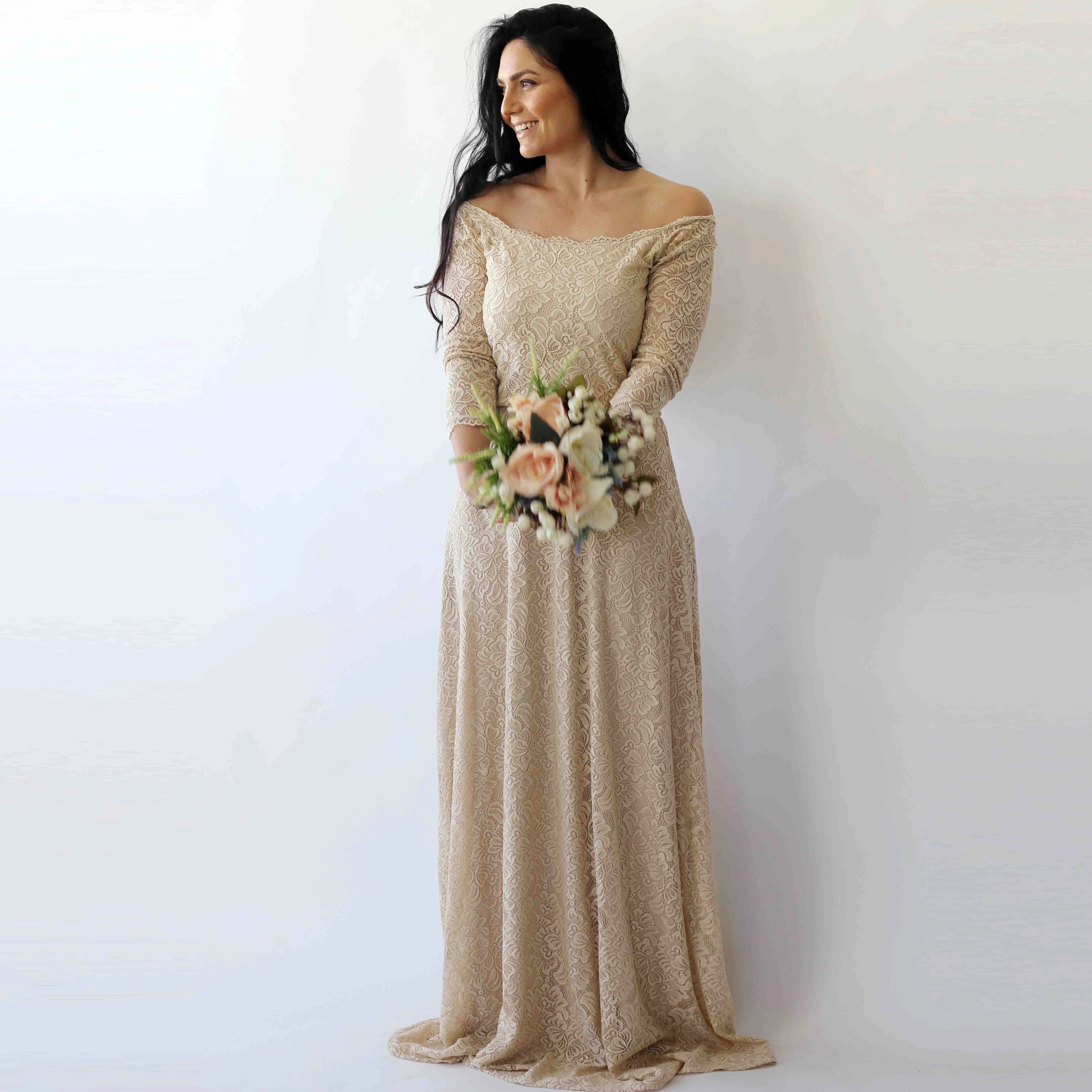 Solenara off-shoulder champagne wedding dress with floral lace, quarter-length sleeves, and A-line silhouette, worn by a model holding flowers.