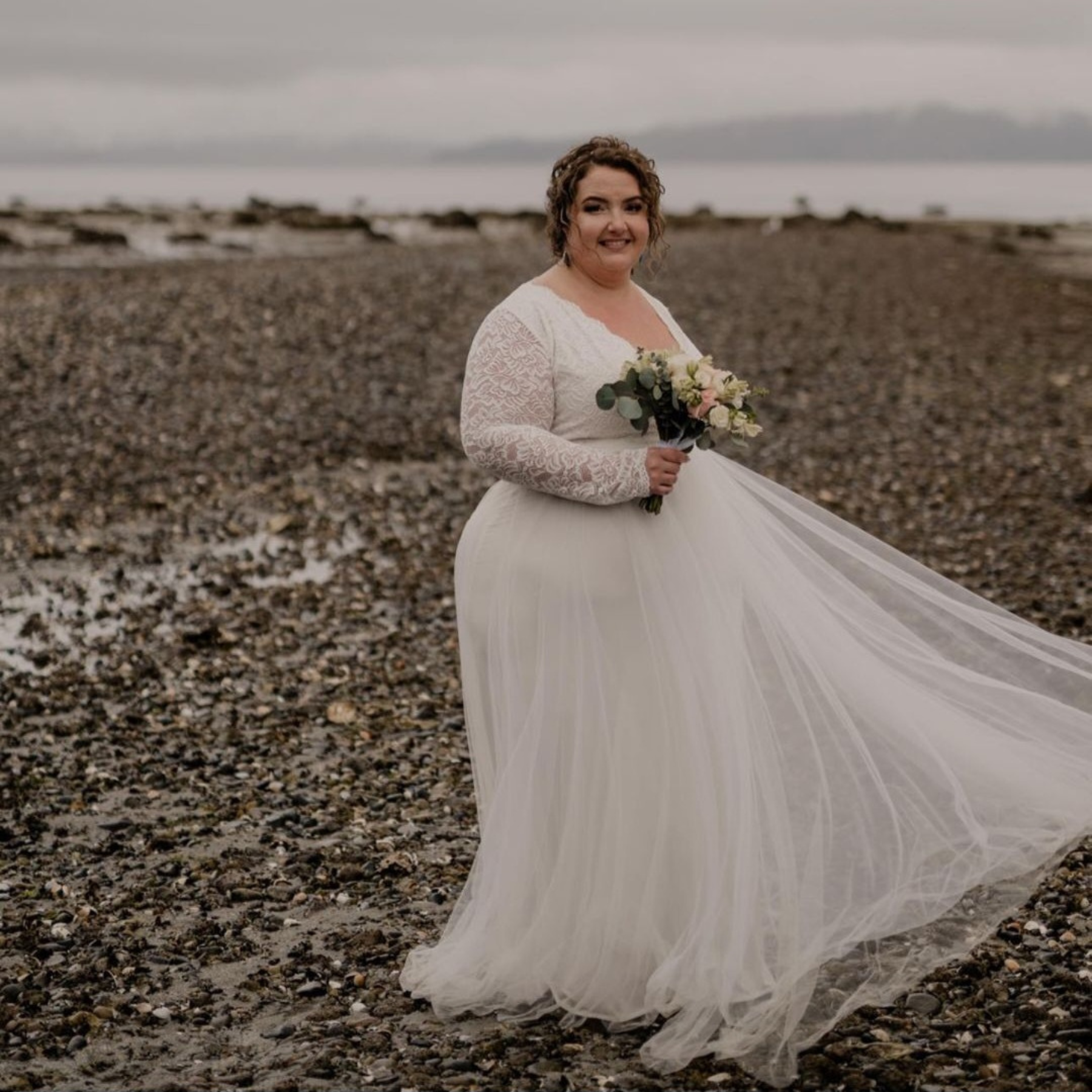Bride in Celestielle Grace Wedding Dress with lace bodice and tulle skirt, holding bouquet on a rocky beach.