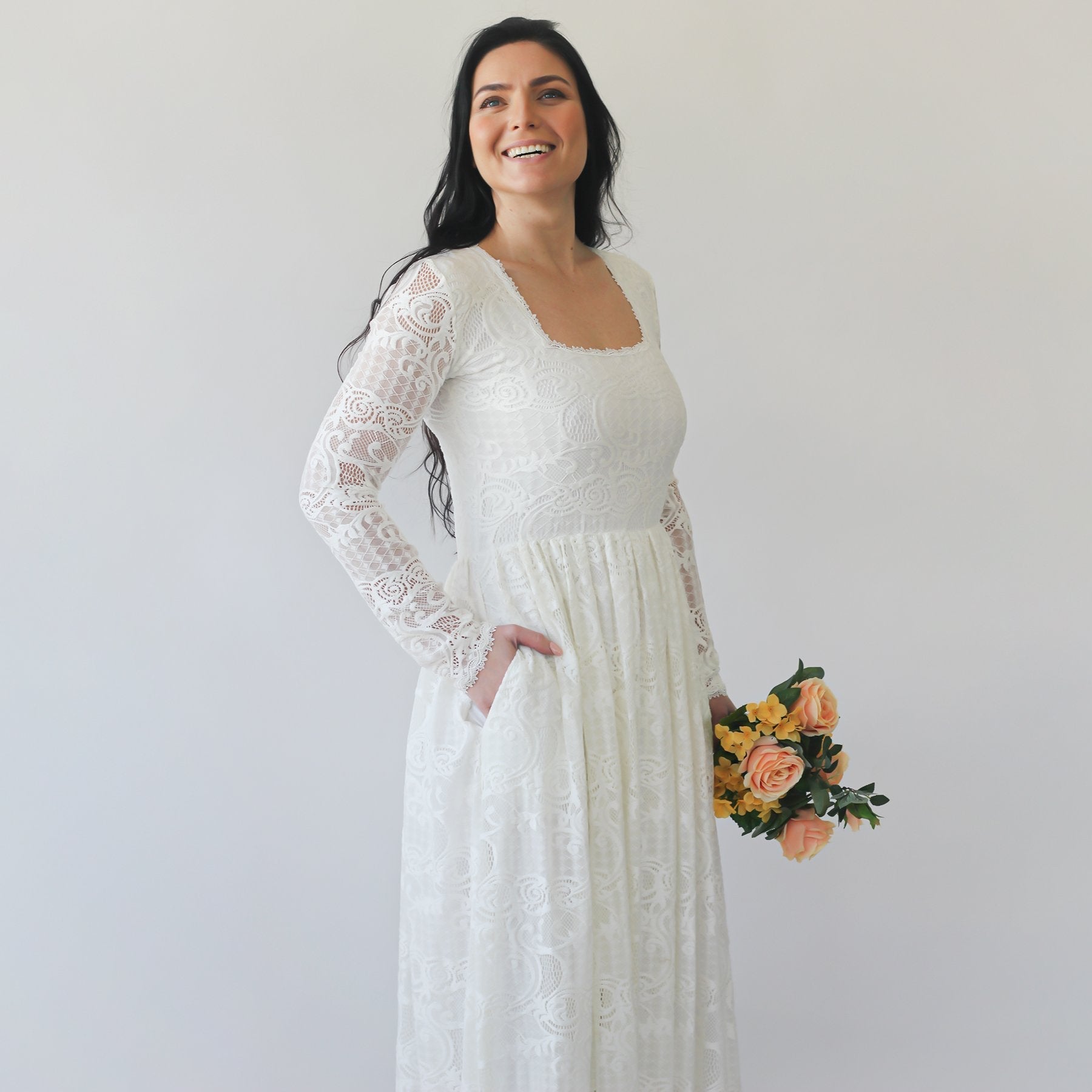 Bride in Nirelda Lace Wedding Dress with square neck and sleeves, holding a bouquet. Ivory gown with floor-length skirt.