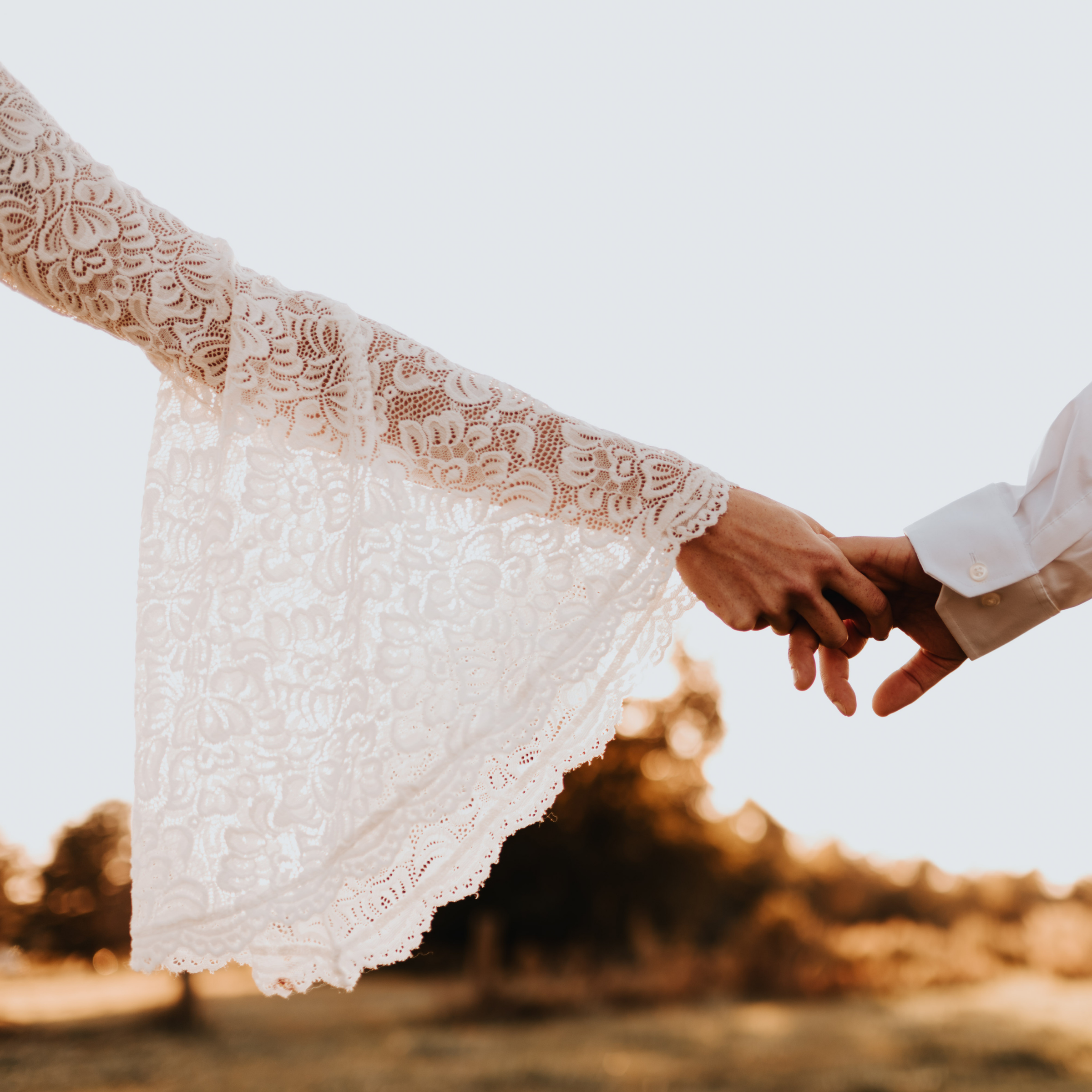 Ivory lace bell sleeve of the Avelyne two-piece wedding ensemble with bride holding groom's hand in outdoor ceremony setting.
