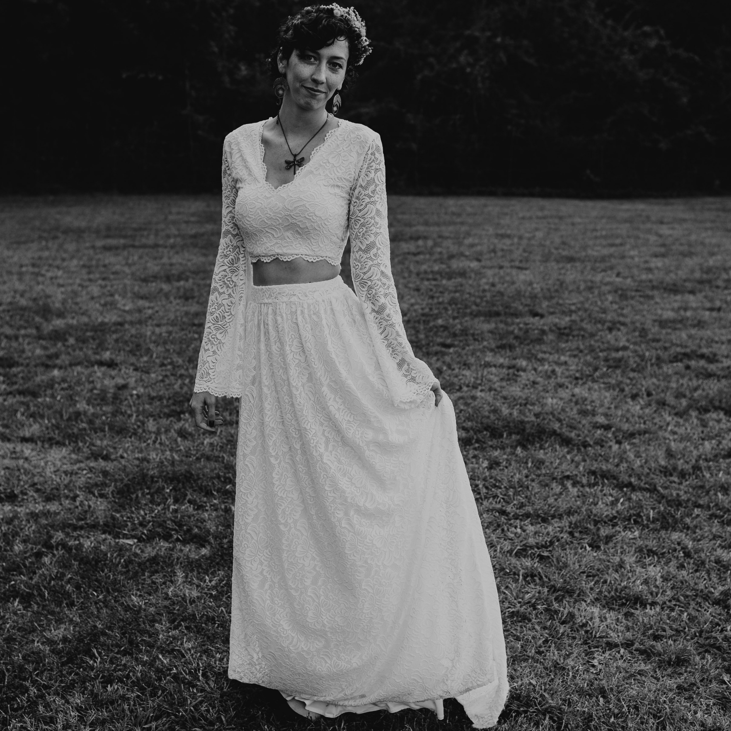 Bride in Avelyne ivory lace two-piece wedding dress with bell sleeves, featuring a flowing skirt and cropped top, in a serene outdoor setting.