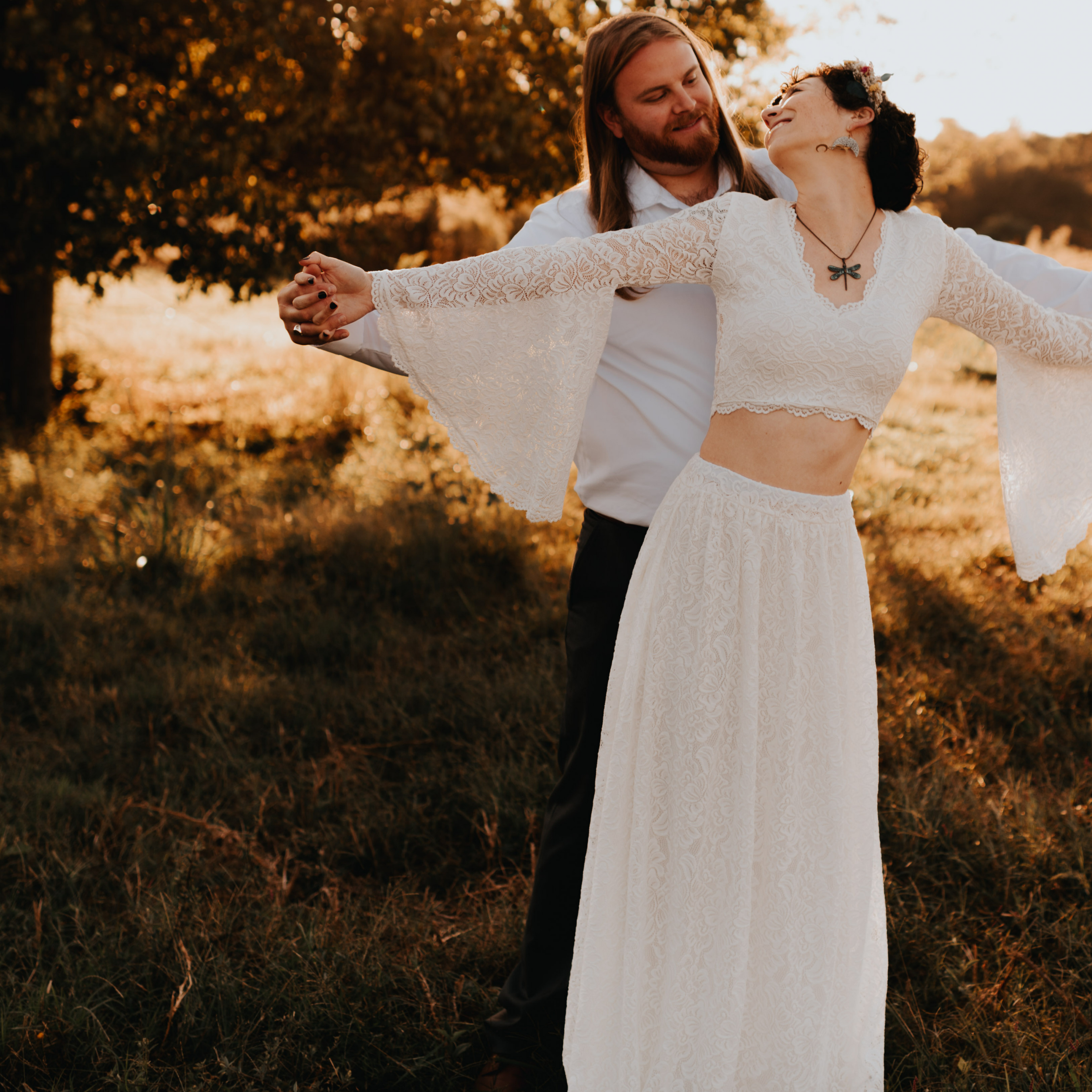Couple in a field with bride wearing an ivory lace two-piece wedding ensemble featuring bell sleeves and a flowing skirt.