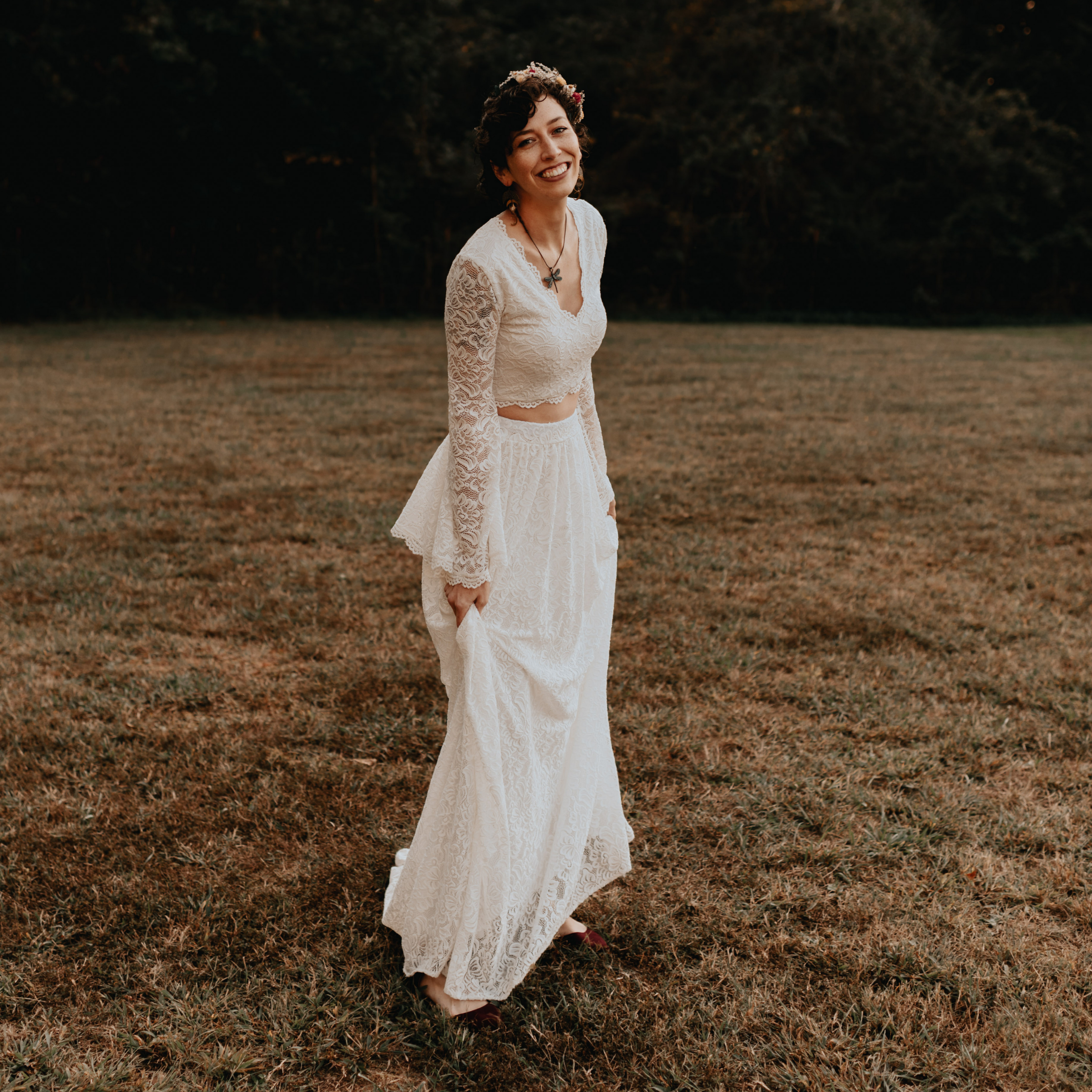 Bride wearing Avelyne ivory lace two-piece wedding ensemble with bell sleeves, standing on grass, showcasing elegance and bohemian grace.