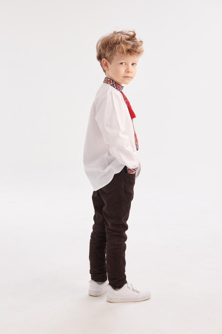 Young boy wearing a traditional white Ukrainian vyshyvanka shirt with red and black embroidery, paired with dark pants and white shoes.