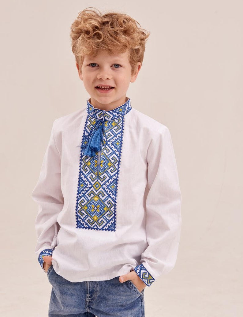 Boy wearing white Ukrainian embroidered shirt with blue and yellow geometric pattern, standing and smiling.