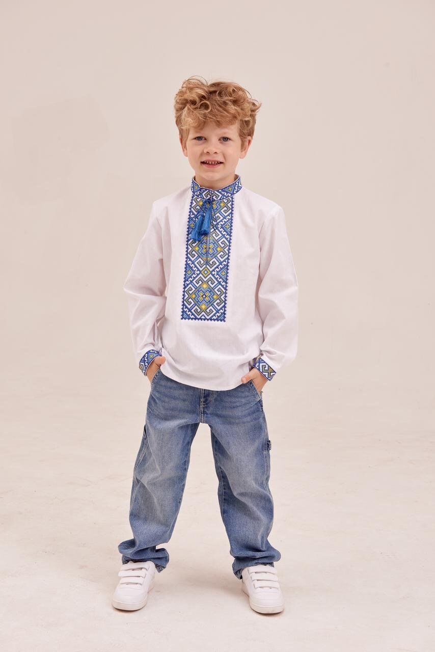 Young boy wearing a traditional white Ukrainian embroidered shirt, vyshyvanka, with blue geometric patterns and tassels.