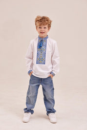Young boy wearing a traditional white Ukrainian embroidered shirt, vyshyvanka, with blue geometric patterns and tassels.