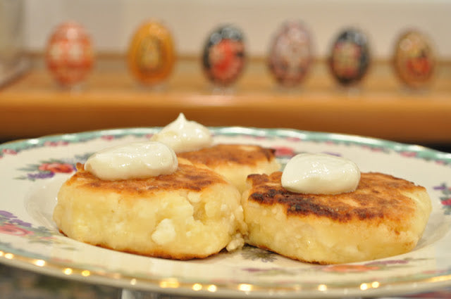 Three golden-brown Ukrainian syrnyky cheese pancakes topped with cream, served on a floral plate.