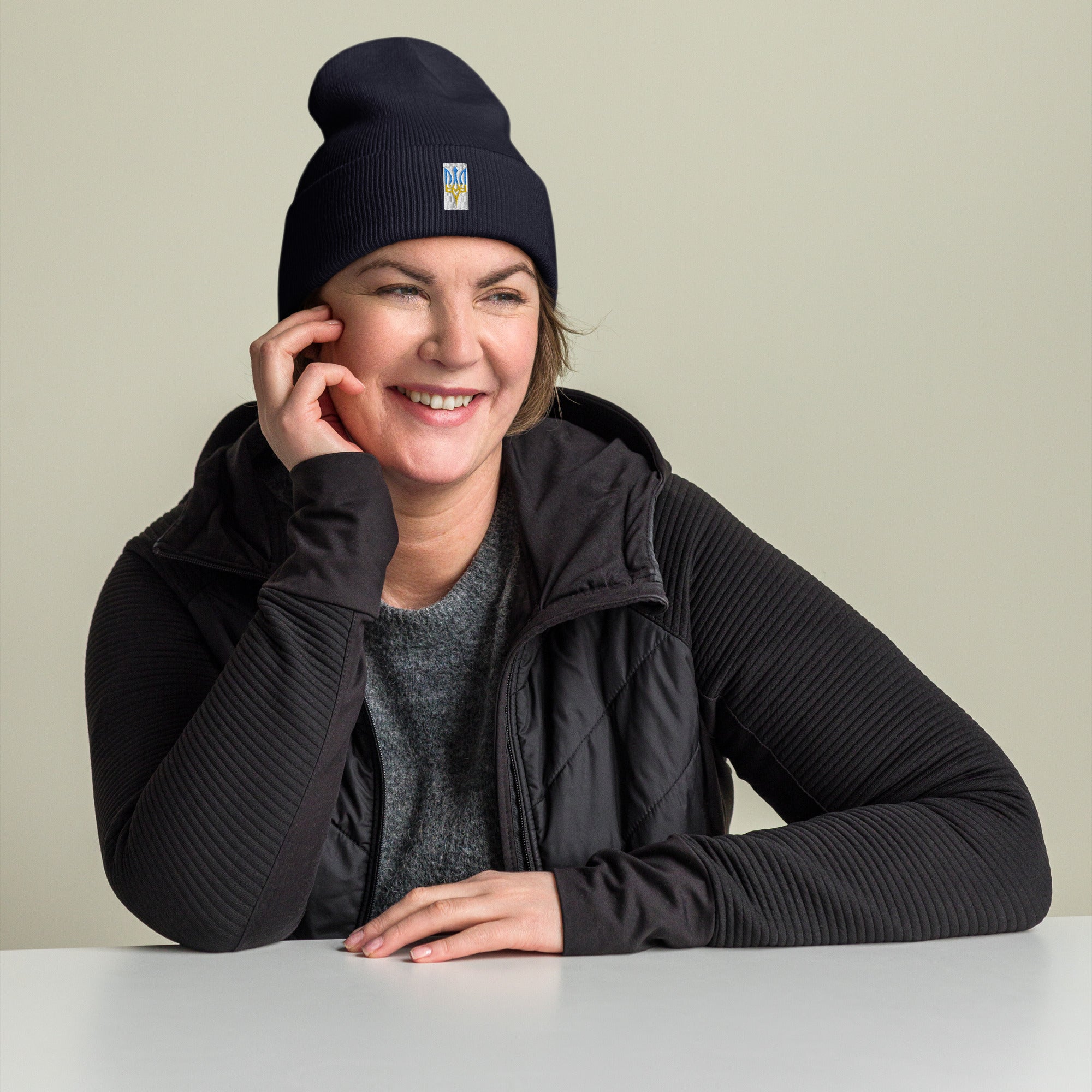 Unisex black cuffed beanie with embroidered Ukrainian Tryzub emblem worn by smiling person in casual outfit.