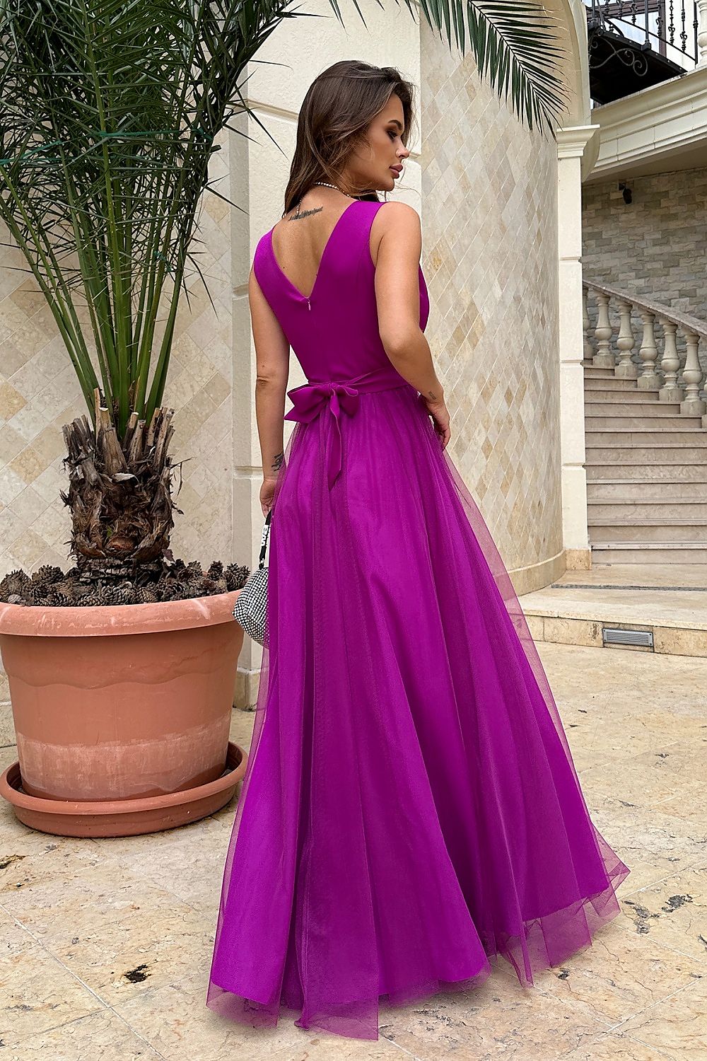 Elegant woman in a striking purple evening dress with a bow, standing outdoors next to a potted palm.