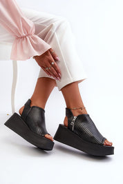 Black openwork leather platform sandals with high heel, worn with white pants, featuring zip closure for casual and refined summer style.