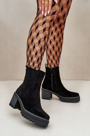 Woman wearing black suede-finish ankle boots with block heel and patterned tights, showcasing stylish footwear in a modern setting.