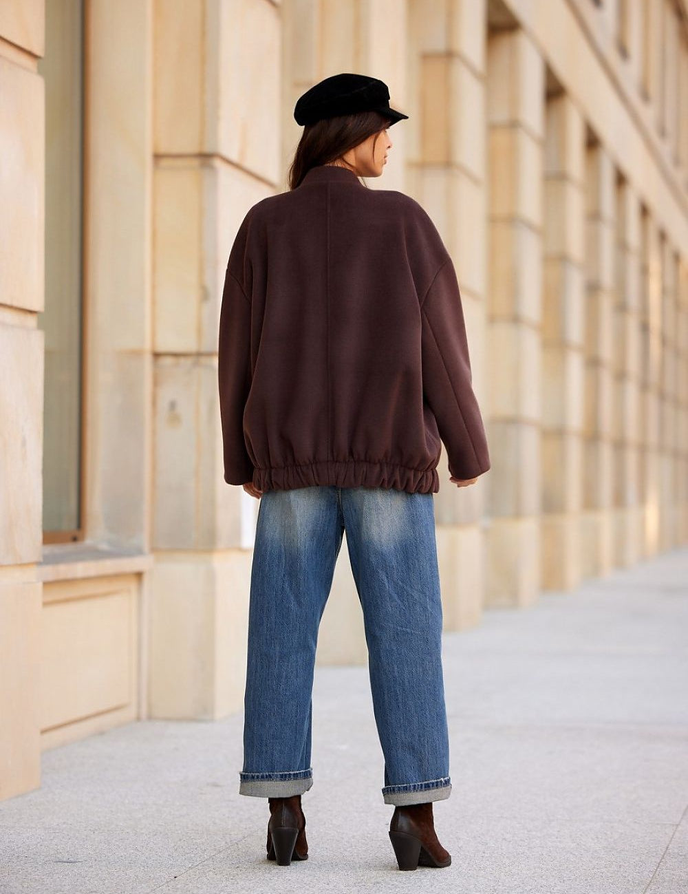 Back view of a woman wearing a Vernelle stand-collar buttoned jacket with dropped shoulders, paired with jeans and a hat.