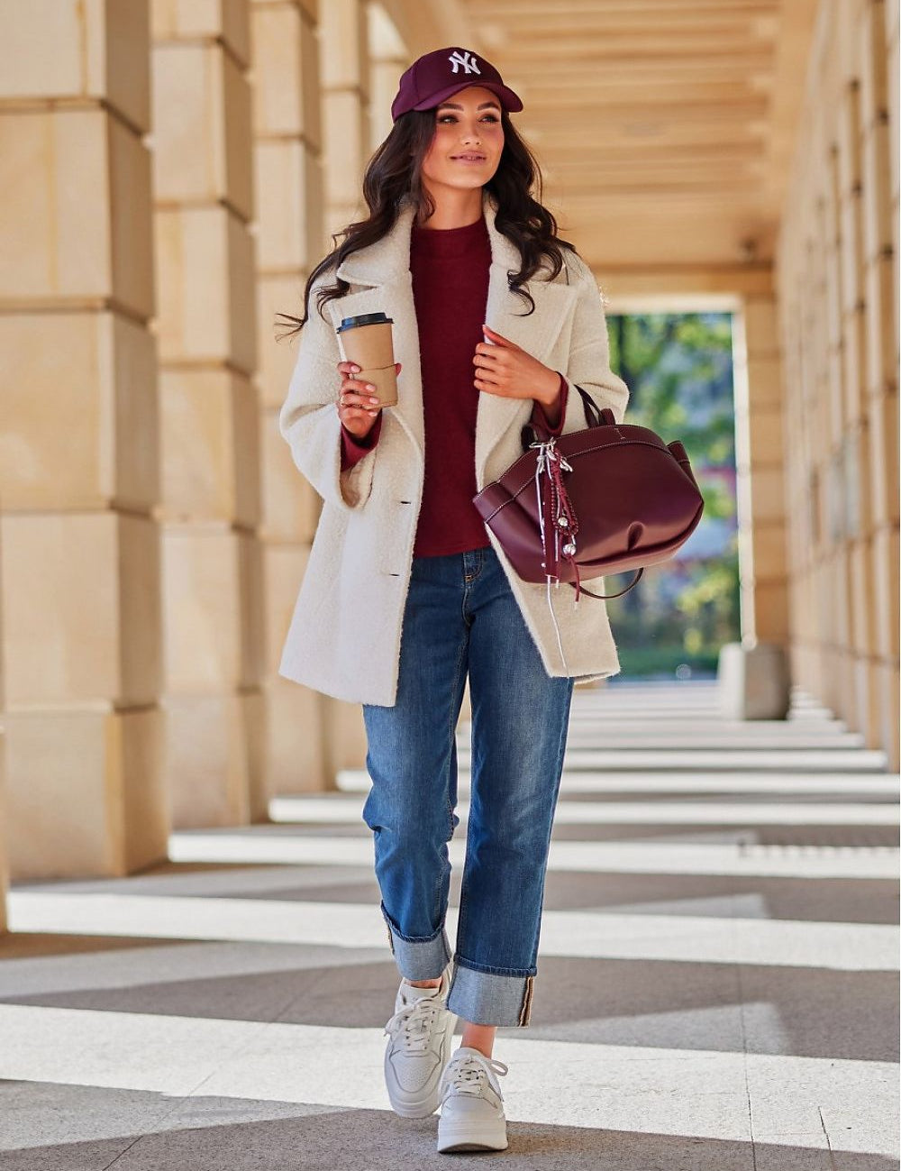 Woman in a white coat walking in a sunlit corridor, holding a coffee and burgundy bag, wearing a matching cap and jeans.