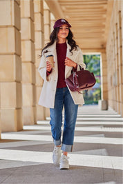 Woman in a white coat walking in a sunlit corridor, holding a coffee and burgundy bag, wearing a matching cap and jeans.