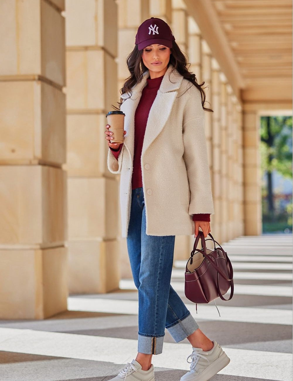 Woman in stylish winter outfit with cream coat, jeans, and sneakers, holding coffee cup, walking under architectural arches.