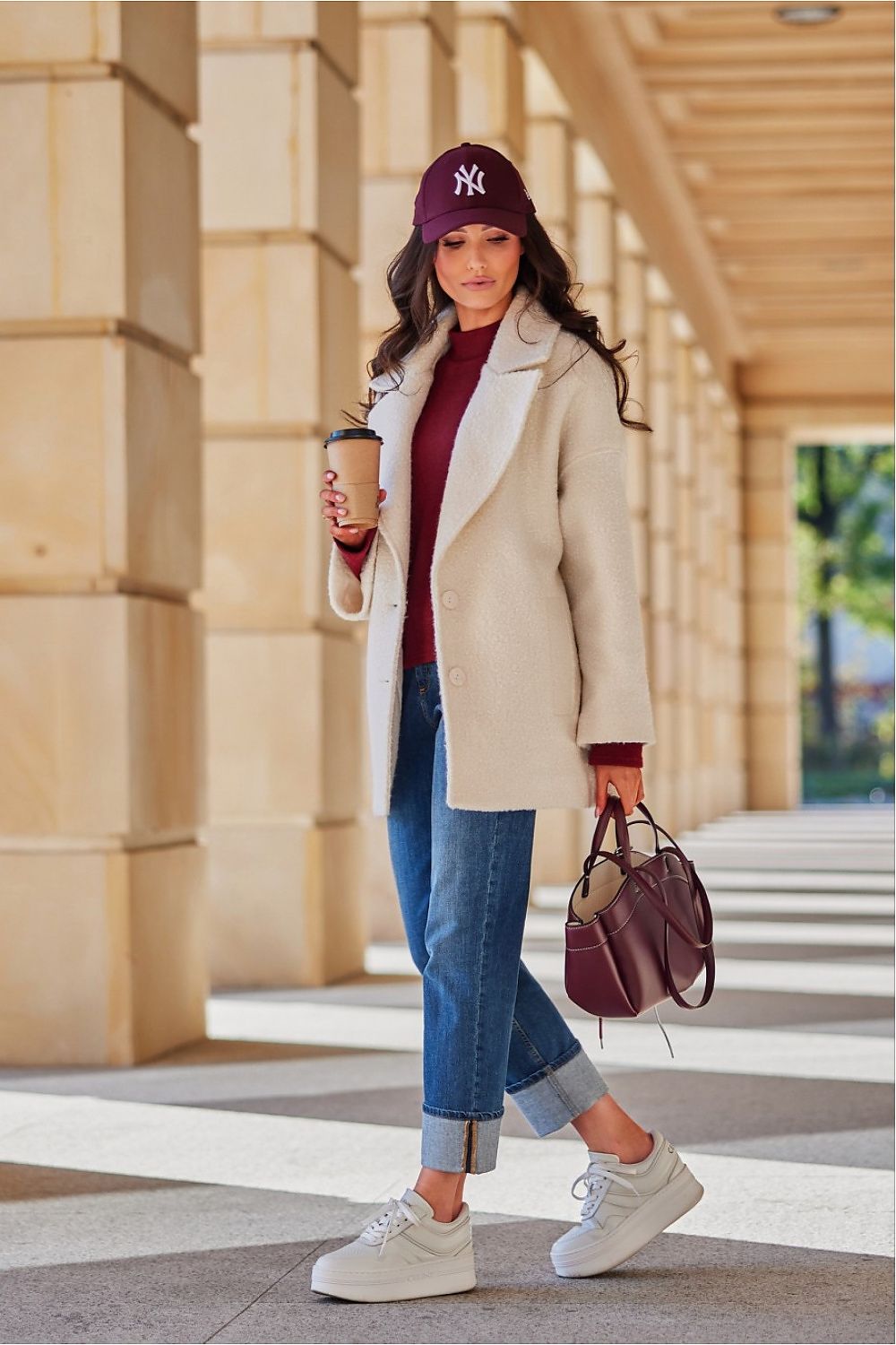 Woman in stylish winter outfit with cream coat, jeans, and sneakers, holding coffee cup, walking under architectural arches.