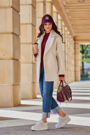 Woman in stylish winter outfit with cream coat, jeans, and sneakers, holding coffee cup, walking under architectural arches.