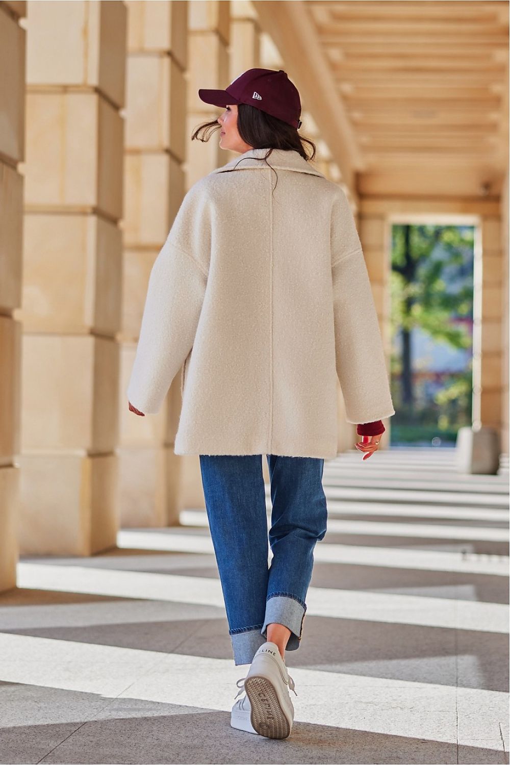 Woman in casual outfit wearing a cream coat and blue jeans walking through a sunlit corridor.