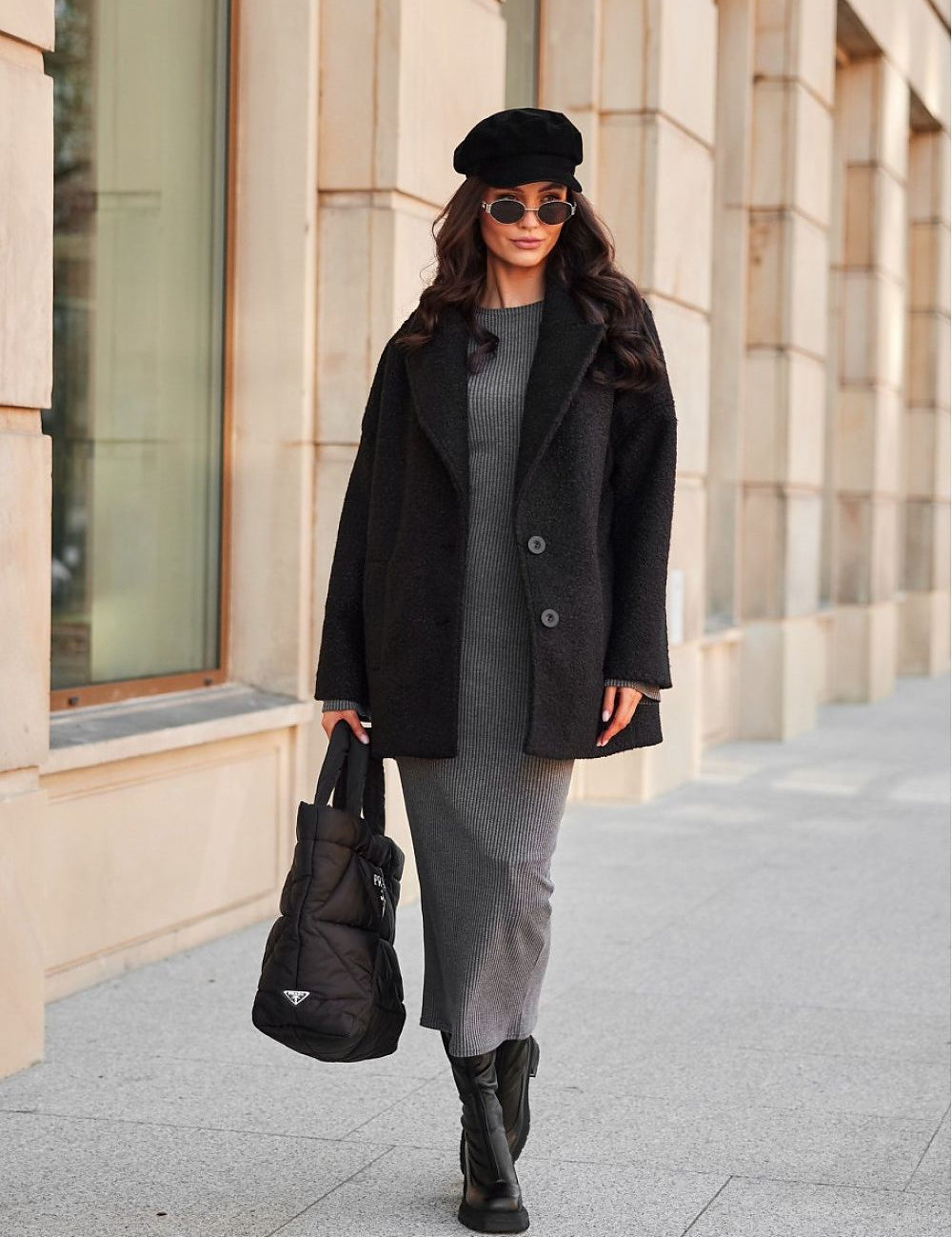 Woman in a black coat and beret, holding a handbag, walking on an urban sidewalk with beige building background.
