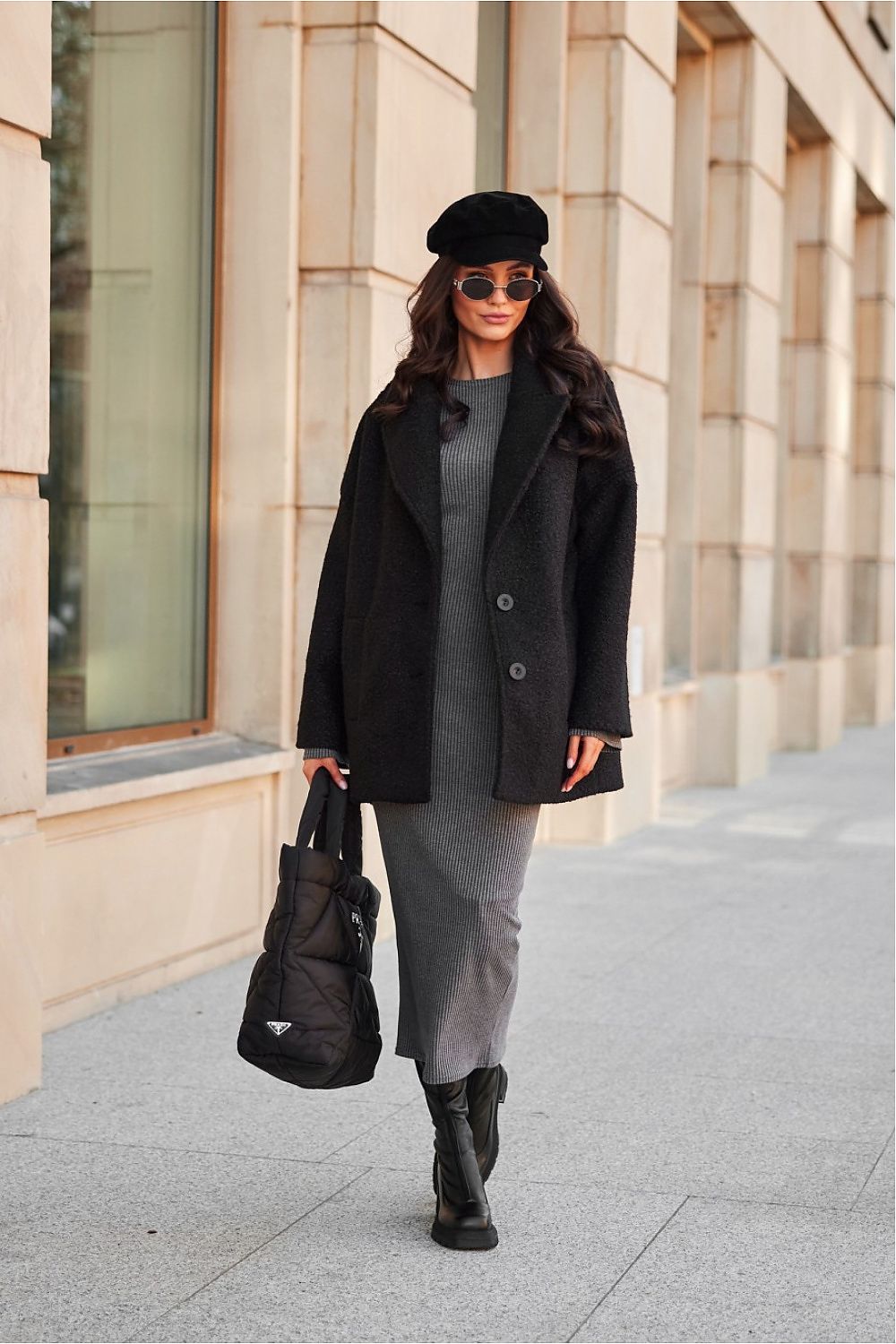 Woman in a black coat and beret, holding a handbag, walking on an urban sidewalk with beige building background.
