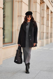 Woman in a black coat and beret, holding a handbag, walking on an urban sidewalk with beige building background.